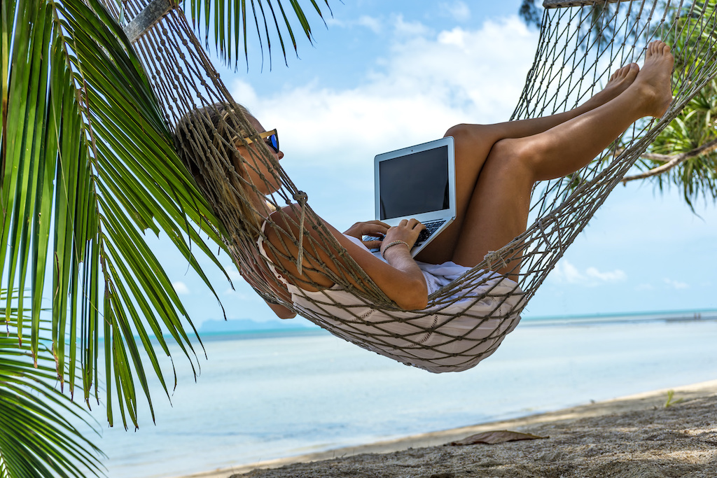 77 Marketing Strategies & Resources for Business Owners - Woman laying in hammock on tropical beach with laptop in her lap, working with the ocean and palm trees